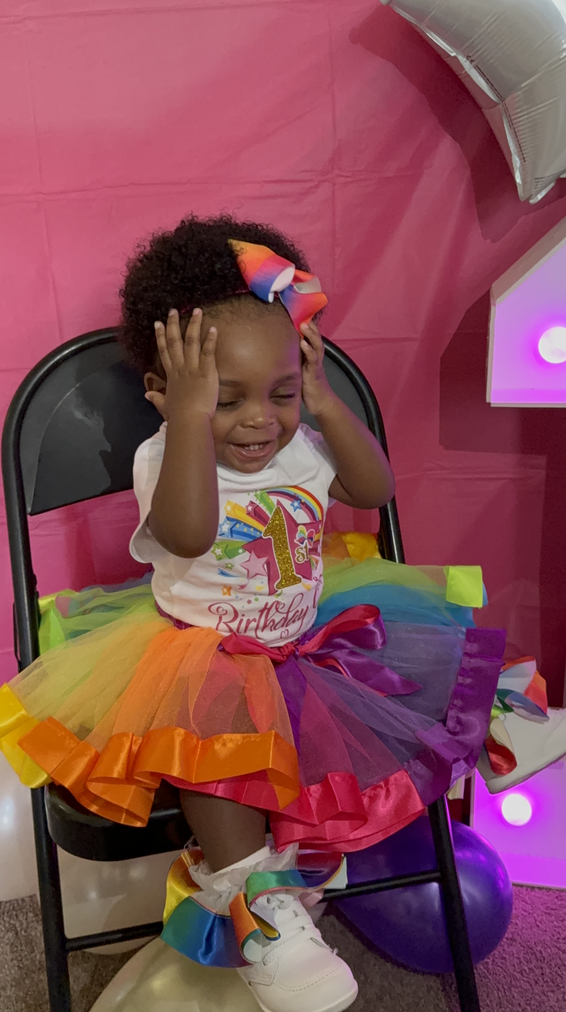 A young girl in a vibrant rainbow tutu and bow sits on a black folding chair, her hands raised to her head as she smiles a...